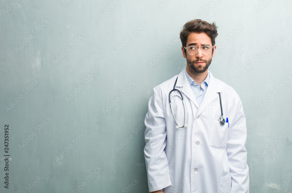 Young friendly doctor man against a grunge wall with a copy space doubting and confused, thinking of an idea or worried about something Young friendly doctor man against a grunge wall with a copy space doubting and confused, thinking of an idea or worried about something