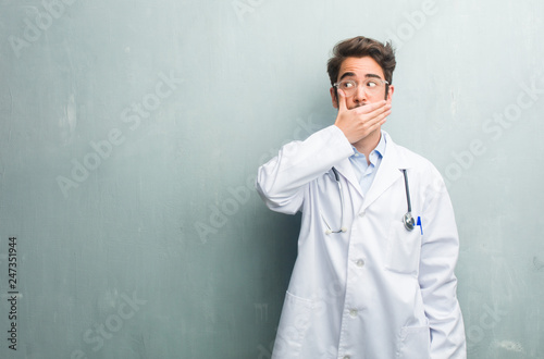Young friendly doctor man against a grunge wall with a copy space covering mouth, symbol of silence and repression, trying not to say anything