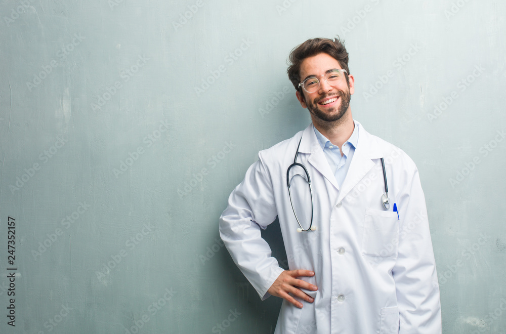Young friendly doctor man against a grunge wall with a copy space with hands on hips, standing, relaxed and smiling, very positive and cheerful Young friendly doctor man against a grunge wall with a copy space with hands on hips, standing, relaxed and smiling, very positive and cheerful