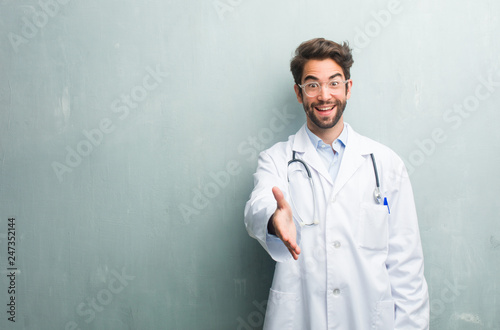 Young friendly doctor man against a grunge wall with a copy space reaching out to greet someone or gesturing to help, happy and excited