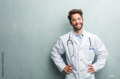 Young friendly doctor man against a grunge wall with a copy space with hands on hips, standing, relaxed and smiling, very positive and cheerful
