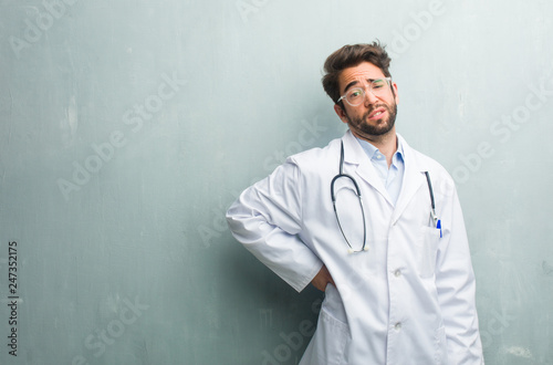 Young friendly doctor man against a grunge wall with a copy space with back pain due to work stress, tired and astute