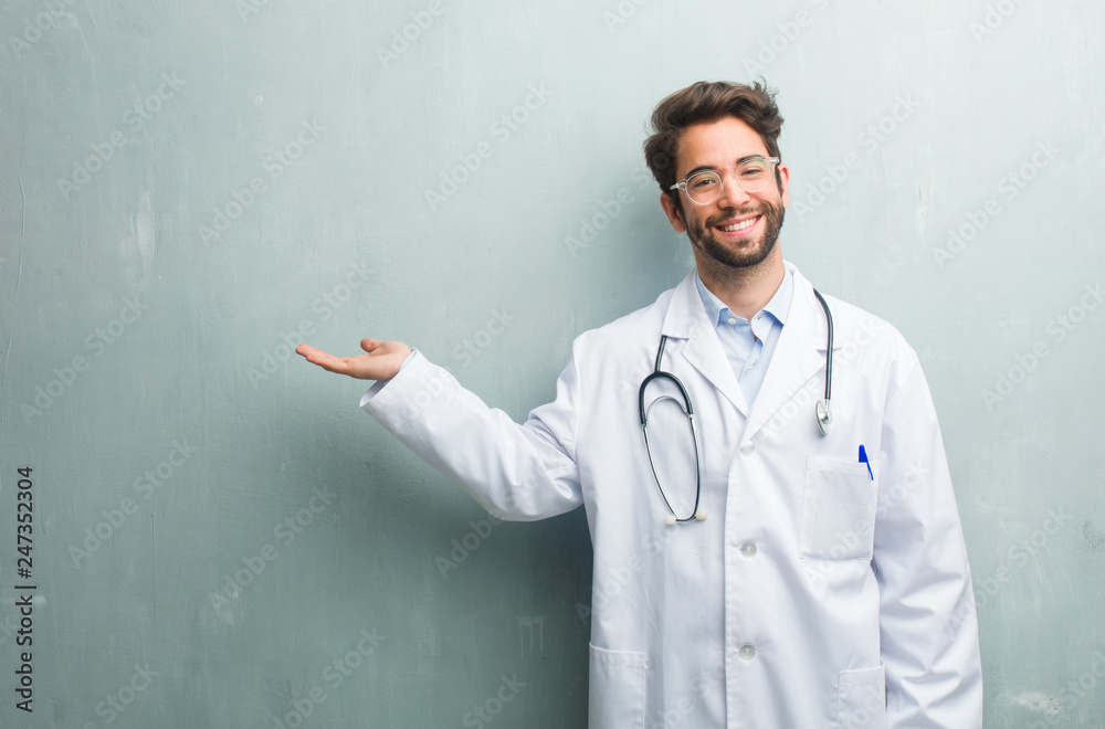 Young friendly doctor man against a grunge wall with a copy space holding something with hands, showing a product, smiling and cheerful, offering an imaginary object Young friendly doctor man against a grunge wall with a copy space holding something with hands, showing a product, smiling and cheerful, offering an imaginary object