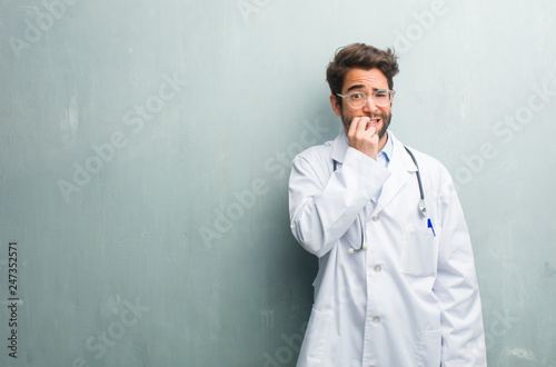 Young friendly doctor man against a grunge wall with a copy space biting nails, nervous and very anxious and scared for the future, feels panic and stress