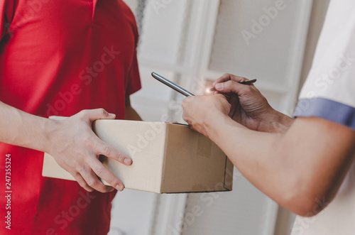 home delivery service man in red uniform and young man customer appending signature in digital mobile phone receiving parcel post box from courier at home, express delivery and online shopping concept