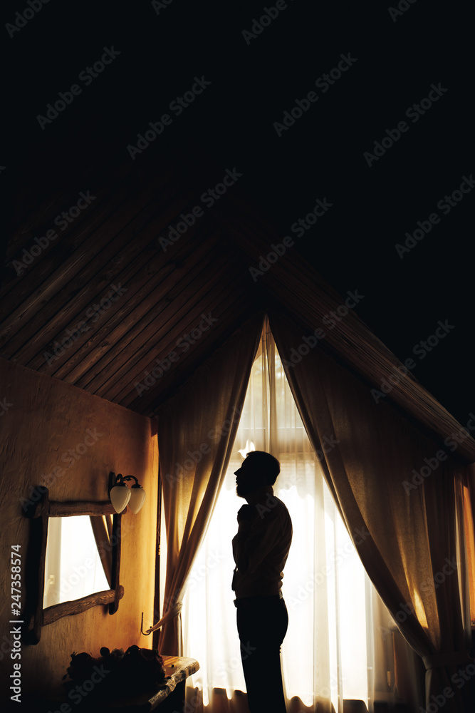 Groom is standing near the window and holding his hands before him. Elegance and attractive photo