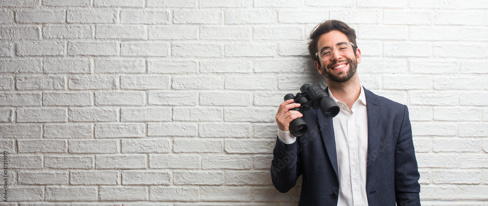 Young business man wearing a suit against a white bricks wall cheerful ...