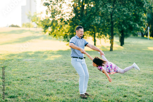 Handsome hipster father with mustache, hair style whirling his little daughter ootdoor on green field in park. Dad playing carousel with his cute child in multicolored dress. Parent with kid enjoying photo