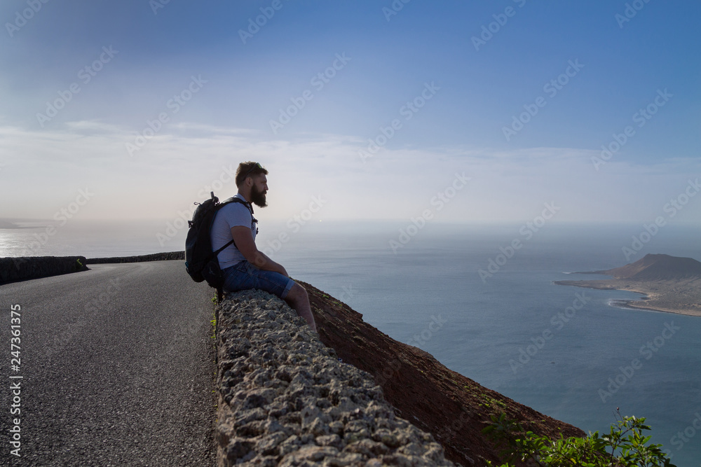 A guy in summer clothes is sitting on a stone fence and looks at a nearby island in the ocean.