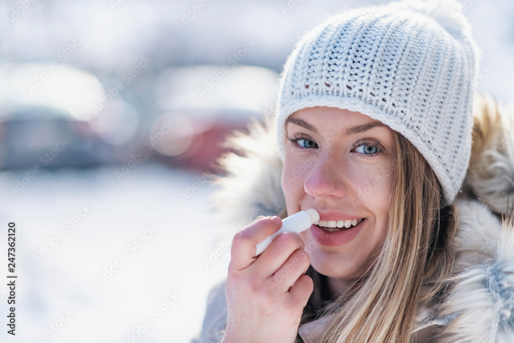 Woman applying lip balsam while walk on a wintery day. - obrazy, fototapety, plakaty 