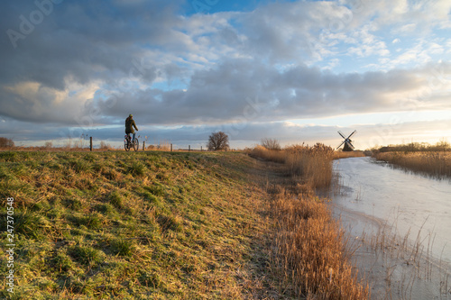 Fotografía Woman cycling in the Dutch countryside near a traditional windmill