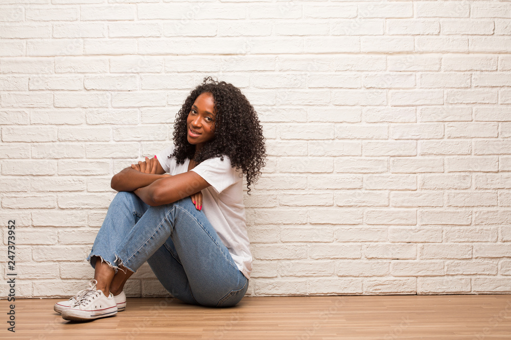 Young black woman sitting on a wooden floor crossing his arms, smiling and happy, being confident and friendly