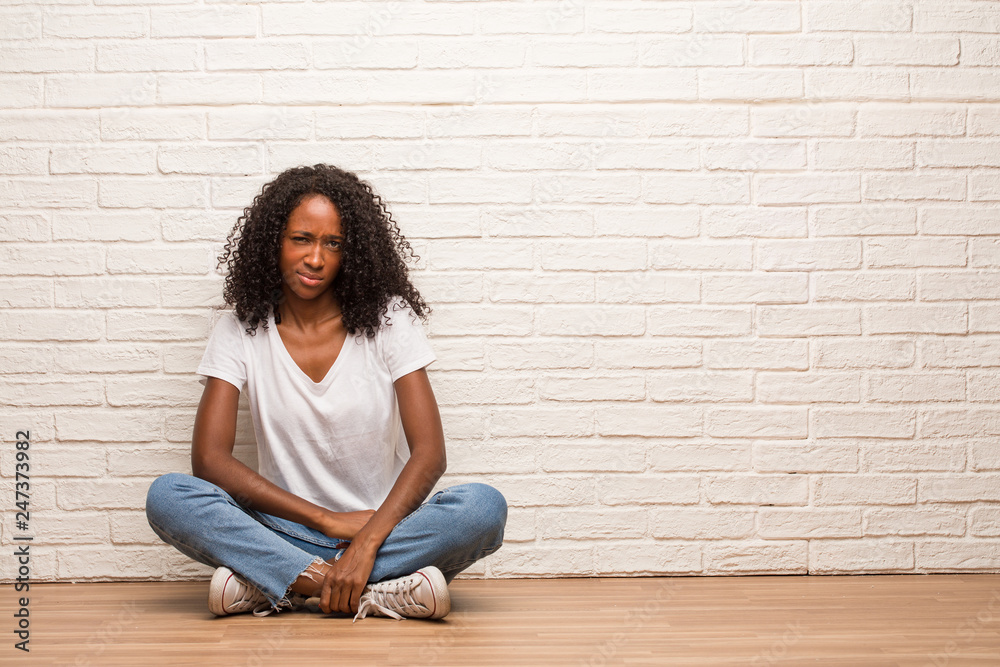 Young black woman sitting on a wooden floor doubting and confused, thinking of an idea or worried about something Young black woman sitting on a wooden floor doubting and confused, thinking of an idea or worried about something