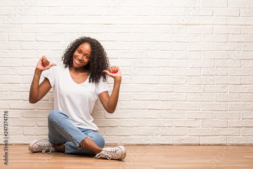 Young black woman sitting on a wooden floor proud and confident, pointing fingers, example to follow, concept of satisfaction, arrogance and health © Asier