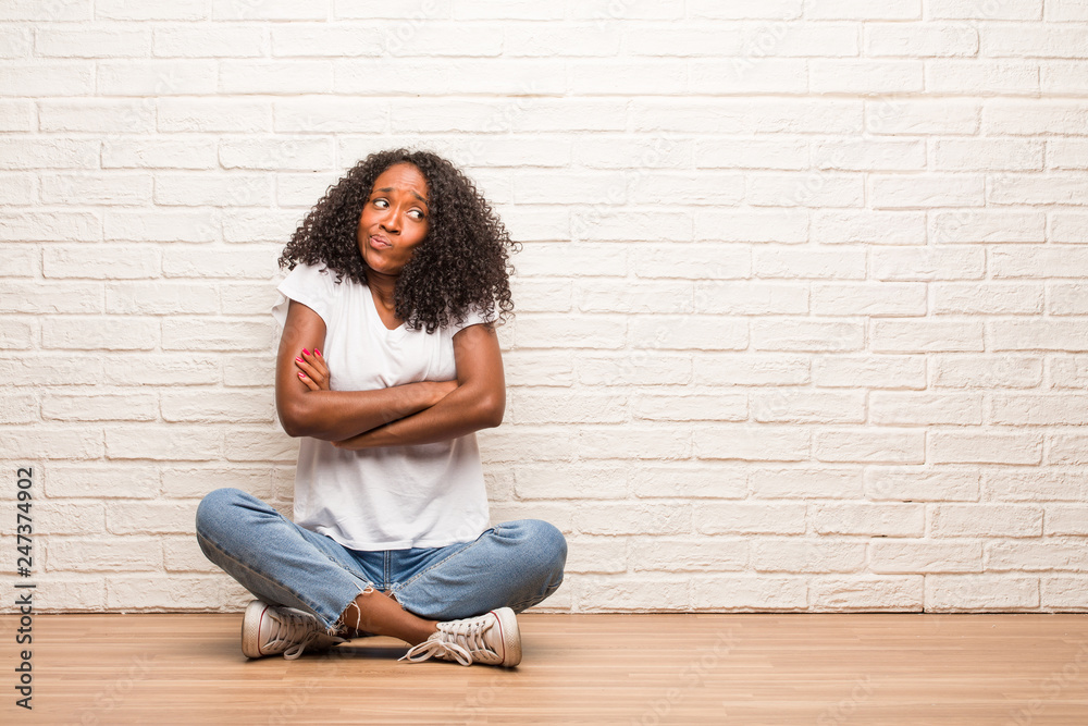 Young black woman sitting on wooden floor doubting and shrugging shoulders, concept of indecision and insecurity, uncertain about something Young black woman sitting on wooden floor doubting and shrugging shoulders, concept of indecision and insecurity, uncertain about something