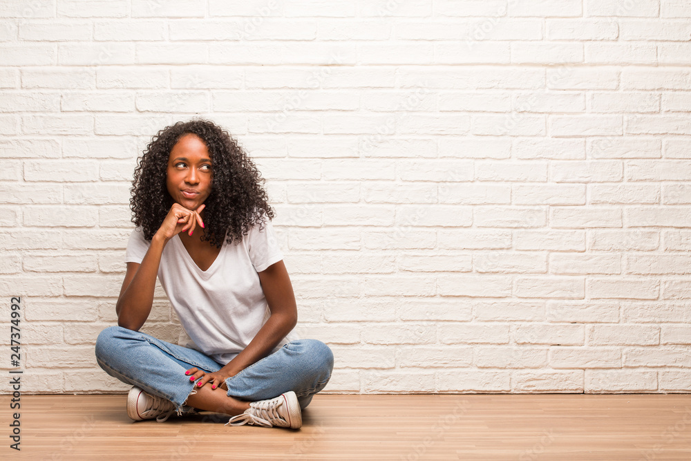 Young black woman sitting on wooden floor thinking and looking up, confused about an idea, would be trying to find a solution Young black woman sitting on wooden floor thinking and looking up, confused about an idea, would be trying to find a solution