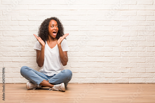 Young black woman sitting on wooden floor surprised and shocked, looking with wide eyes, excited by an offer or by a new job, win concept