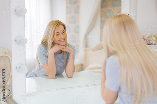 Young attractive woman with long blonde hair and mask under eyes looking to the mirror. Beautiful female with natural makeup  have golden collagen patches on fresh facial skin in the bedroom. 