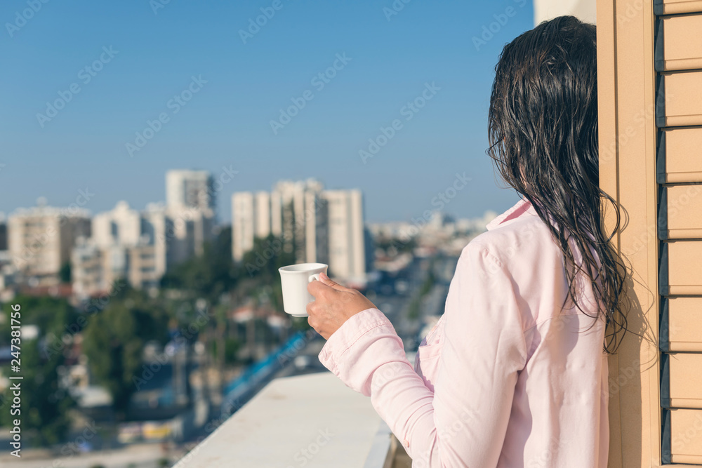 Fototapeta premium Woman with wet hear in a pink shirt with cup of coffee at the summer morning against the background of town