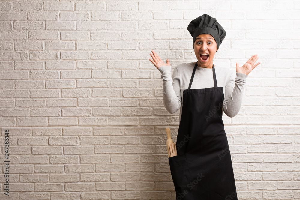 Young baker indian woman against a bricks wall screaming happy, surprised by an offer or a promotion, gaping, jumping and proud