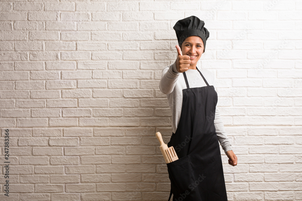 Young baker indian woman against a bricks wall cheerful and excited, smiling and raising her thumb up, concept of success and approval, ok gesture Young baker indian woman against a bricks wall cheerful and excited, smiling and raising her thumb up, concept of success and approval, ok gesture
