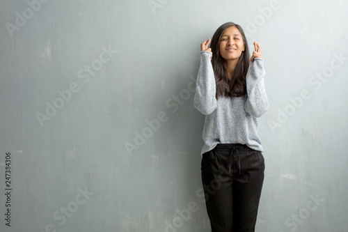 Young indian woman against a grunge wall crossing his fingers, wishes to be lucky for future projects, excited but worried, nervous expression closing eyes