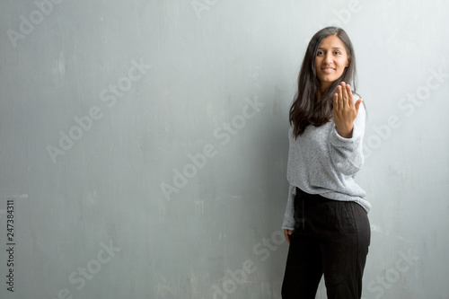 Young indian woman against a grunge wall inviting to come, confident and smiling making a gesture with hand, being positive and friendly