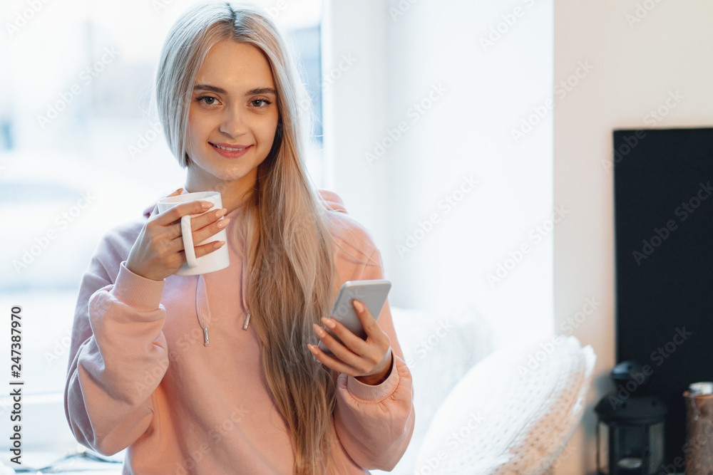 Pretty young woman enjoying coffee while using a smartphone by the windows indoors Pretty young woman enjoying coffee while using a smartphone by the windows indoors