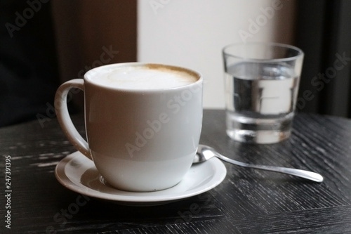 Latte coffee in a white cup on old wooden table with glass of water and spoon.