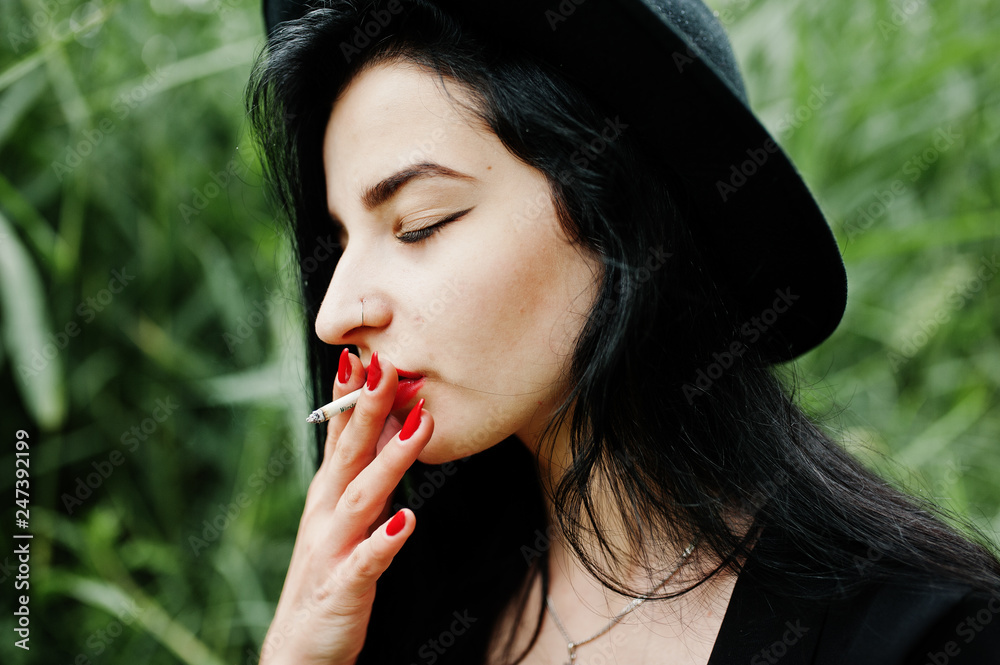 Sensual smoker girl all in black, red lips and hat. Goth dramatic woman smoking on common reed. Sensual smoker girl all in black, red lips and hat. Goth dramatic woman smoking on common reed.
