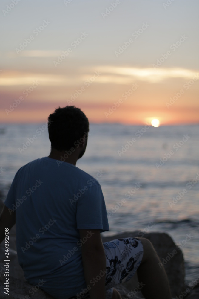 Young man sitting alone on the seashore during sunset. Warm, pleasant, summer evening on the Croatian coast. Calm end of the day on the shore. Vacation, rest in Croatia. Dusk, twilight, Adriatic Sea