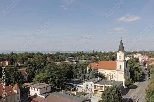 A view of the city from the water tower in Siofok. Visible buildings, church, streets. Lake Balaton in the background. A warm, nice, sunny, calm summer day. White clouds, blue sky. Holidays in Hungary