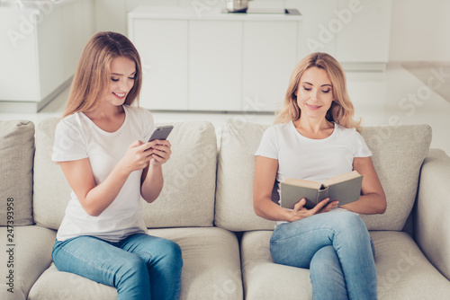 Two nice adorable attractive charming lovely winsome cute ladies sitting on divan using gadget reading having free spare time in light white interior room indoors