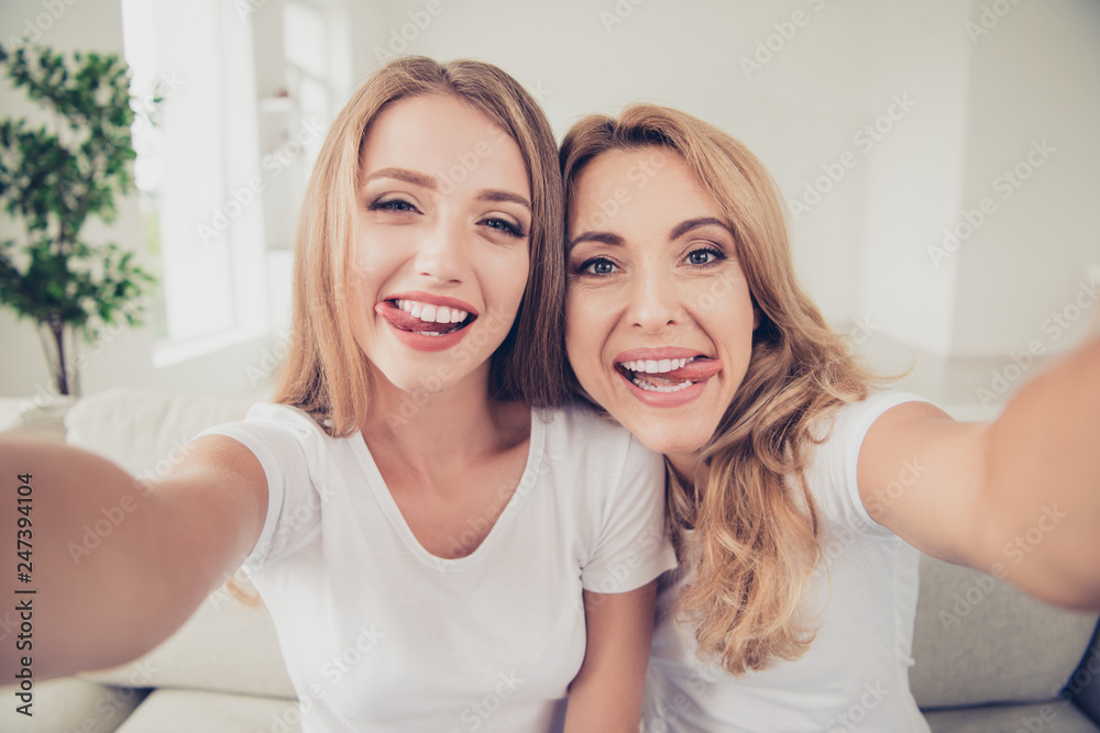 Close up photo of two people mum and teen daughter holding hands arms telephone funny tongue out of mouth make take selfie for dad daddy father wear white t-shirts jeans sit on comfy sofa Close up photo of two people mum and teen daughter holding hands arms telephone funny tongue out of mouth make take selfie for dad daddy father wear white t-shirts jeans sit on comfy sofa