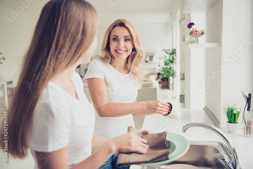 Close up photo two people mum and teen daughter enjoy spending time help with house work duties wash dry plates overjoyed best company wear white t-shirts jeans in bright flat kitchen
