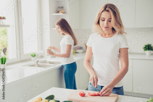 Two nice lovely attractive charming cheerful people blonde mom mum cooking tasty food girl cleaning kitchenware in modern light white interior room