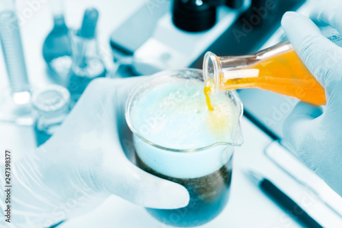 Close up of chemist hand pouring yellow liquid oil in beger glass to erlenmeyer flask chemistry with blue liquid on biochemical experiment. Mix water in laboratory.