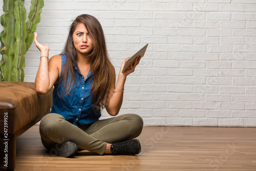 Portrait of young latin woman sitting on the floor crazy and desperate, screaming out of control, funny lunatic expressing freedom and wild. Holding a tablet.