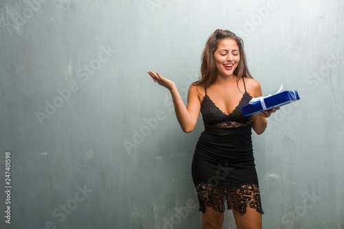 Portrait of young pretty woman wearing a dress against a wall laughing and having fun, being relaxed and cheerful, feels confident and successful. Holding a gift. © Asier