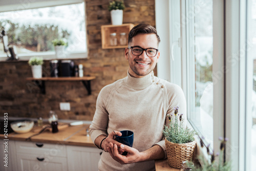 Portrait of smiling millenial man drinking tea near the window at cozy home on winter morning. photo