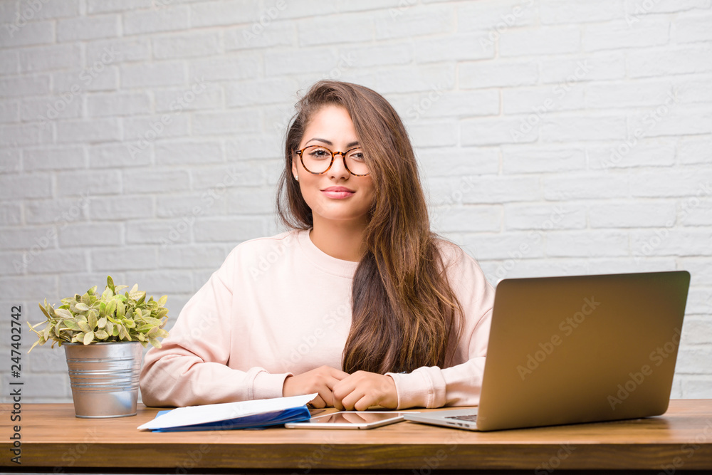 Portrait of young student latin woman sitting on her desk cheerful and with a big smile, confident, friendly and sincere, expressing positivity and success Portrait of young student latin woman sitting on her desk cheerful and with a big smile, confident, friendly and sincere, expressing positivity and success