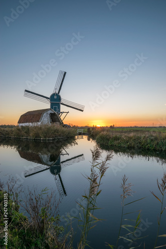 Dutch windmill and house in decay at sunset. Tranquil scene with a beautiful serene sky and reflections of the sky and mill in the water of the canal.