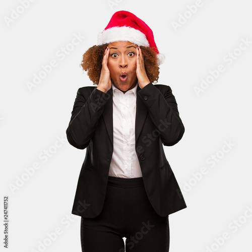 Young business black woman wearing a chirstmas santa hat surprised and shocked