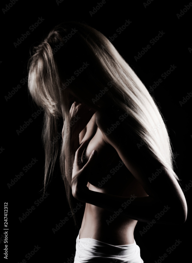 Beautiful slim topless blonde girl with hair falling on her face, stands  sideways with a her hand covering her big boobs. Black background. Artistic  noir silhouette photo Stock Photo