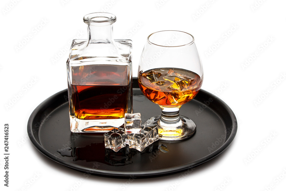 A glass of whiskey with ice or brandy and a square carafe on a black dish isolated against a white background. Whiskey with ice in the glass on a white background. A glass of whiskey with ice or brandy and a square carafe on a black dish isolated against a white background. Whiskey with ice in the glass on a white background.