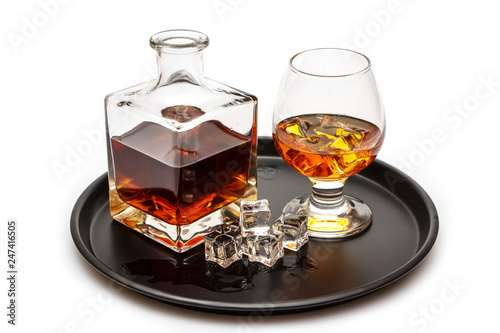 A glass of whiskey with ice or brandy and a square carafe on a black dish isolated against a white background. Whiskey with ice in the glass on a white background.