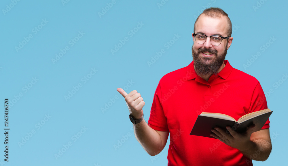 Young hipster man wearing glasses and reading a book over isolated background pointing and showing with thumb up to the side with happy face smiling Young hipster man wearing glasses and reading a book over isolated background pointing and showing with thumb up to the side with happy face smiling