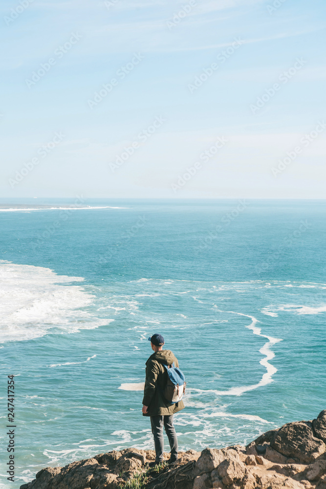 A man tourist with a backpack stands in solitude at Cape Roca in Portugal and enjoys a beautiful view of the Atlantic Ocean.