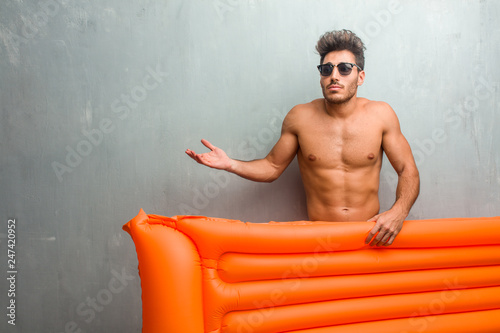 Young athletic man wearing a swimsuit against a grunge wall doubting and shrugging shoulders, concept of indecision and insecurity, uncertain about something