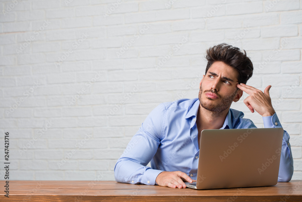 Young business man sitting and working on a laptop making a suicide gesture, feeling sad and scared forming a gun with fingers Young business man sitting and working on a laptop making a suicide gesture, feeling sad and scared forming a gun with fingers